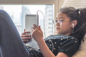 Let's Keep Kids Safe on Social Media, young girl on a tablet with headphones while laying in bed